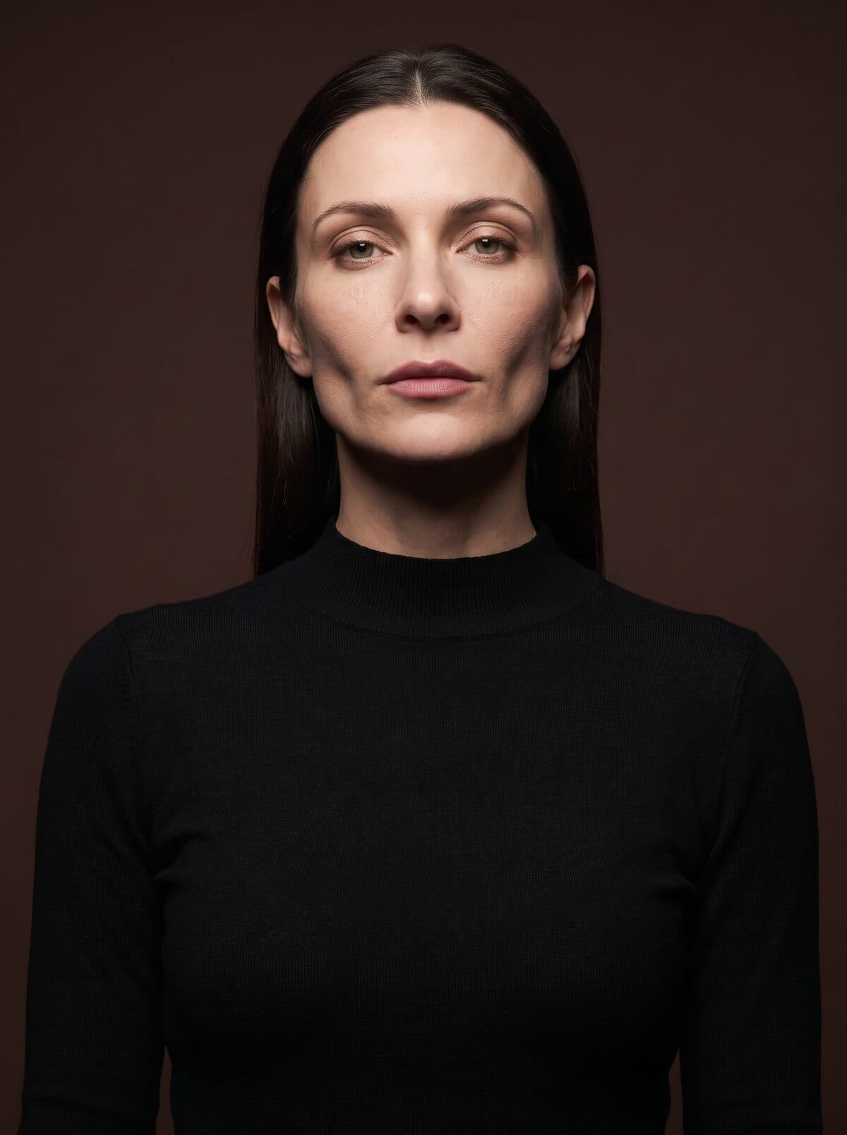 A woman with long dark hair in a black roll-neck, against a deep brown backdrop