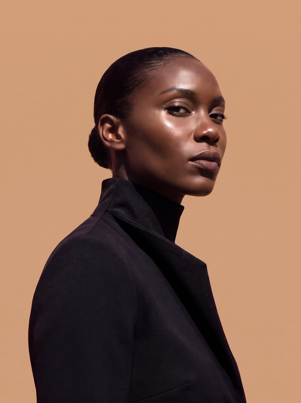 A woman in a black coat and roll-neck, turned to the side against a warm sand backdrop