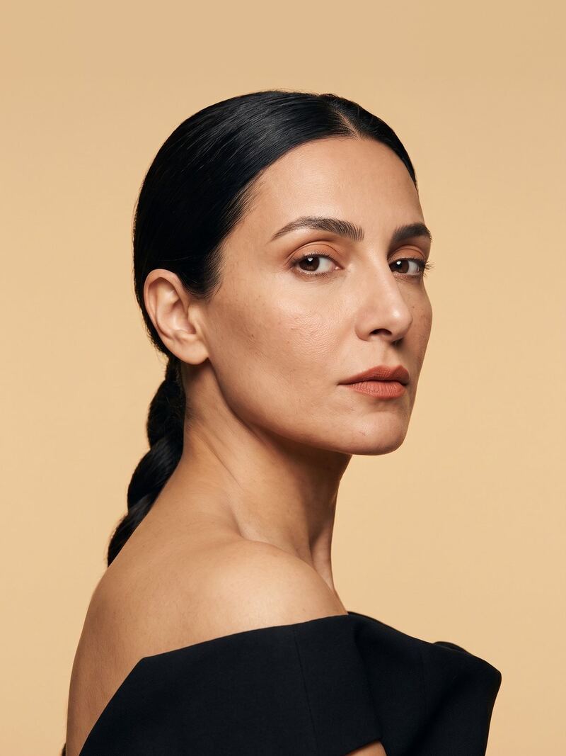 A woman with dark hair in a low braid, looking over her shoulder, against a pale sand backdrop