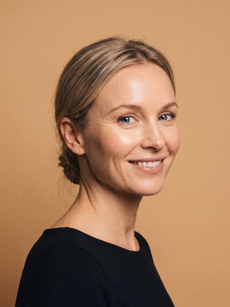 A smiling woman with fair hair pulled back, in a black top, against a warm tan backdrop