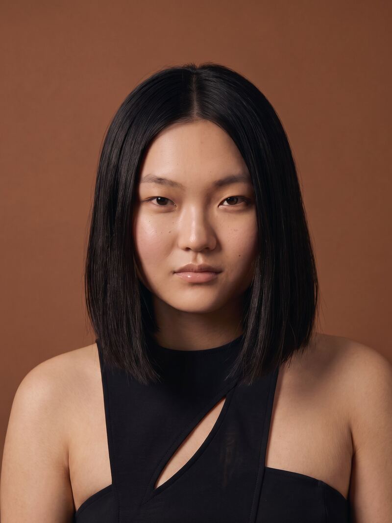 A woman with a straight black bob in a black top, against a warm brown backdrop